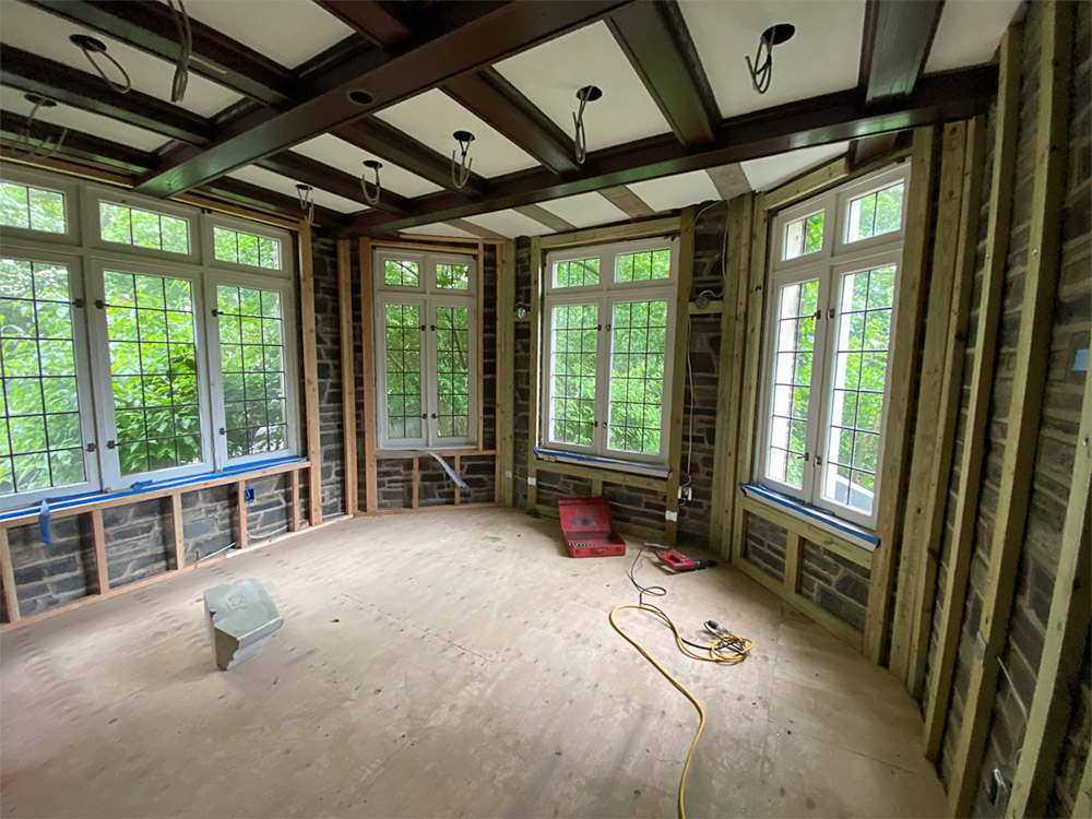 Room under renovation with exposed framing, large grid-style windows letting in natural light, coffered ceiling in progress, and electrical wiring hanging down.