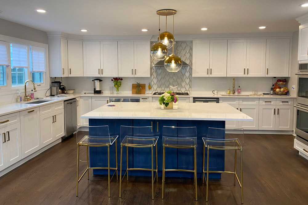 This stunning kitchen renovation showcases a perfect balance of modern elegance and bold design choices. The white shaker cabinetry paired with a vibrant navy-blue island creates a striking contrast, while the gold accents in the hardware, lighting, and barstools add a touch of luxury.