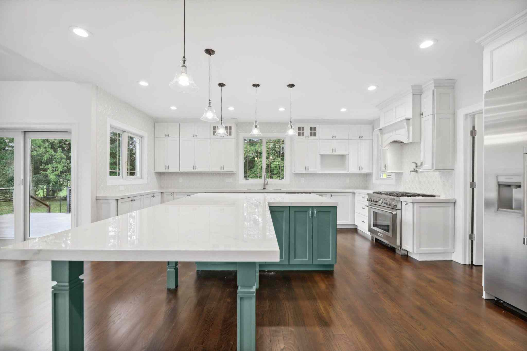 Spacious white kitchen with a large island featuring teal cabinetry, pendant lighting, stainless steel appliances, and dark hardwood floors. Surrounded by windows and sliding glass doors.