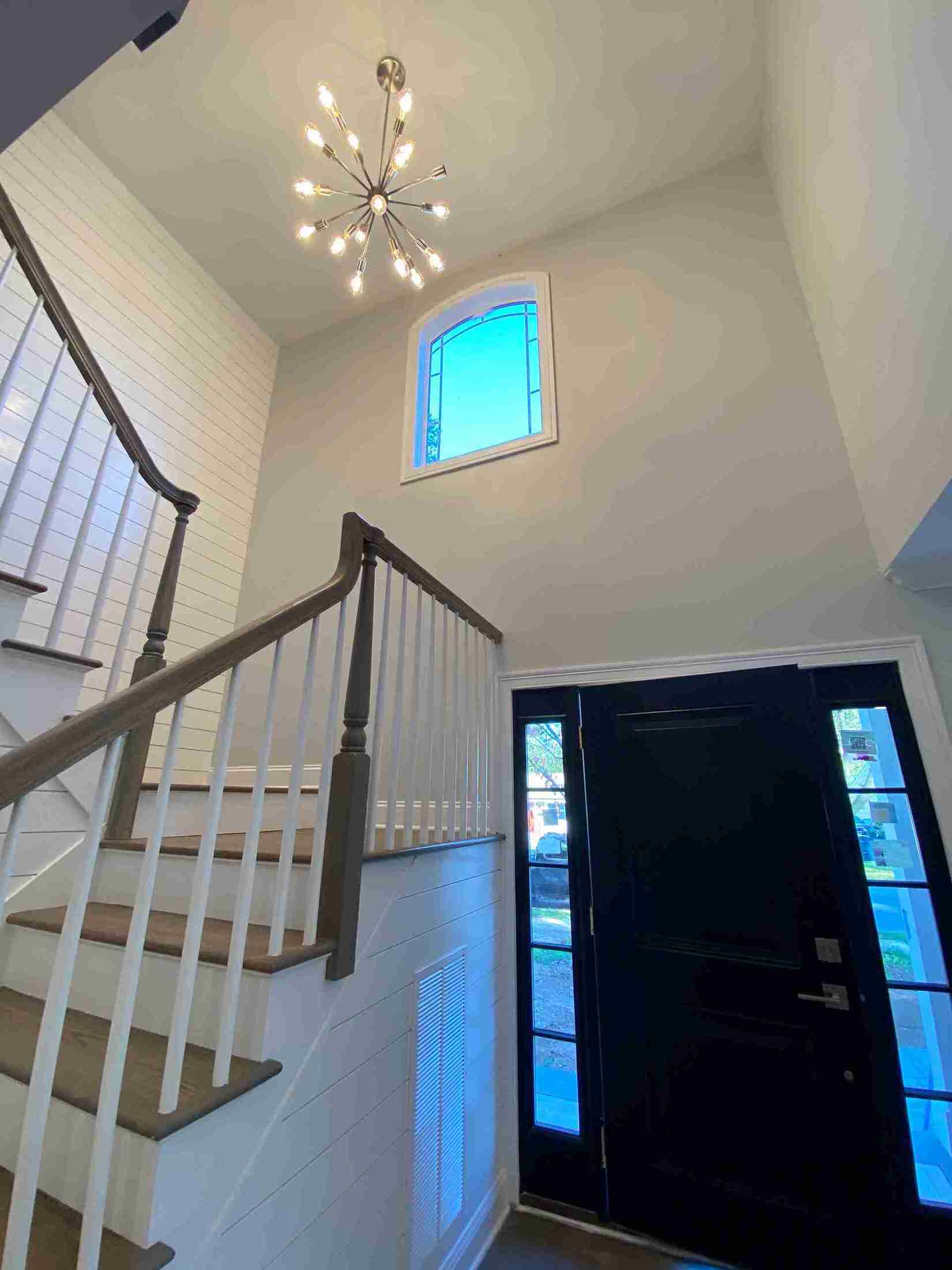 Bright foyer with a modern sputnik chandelier, dark front door with glass panels, wood staircase, shiplap walls, and tall arched window above.