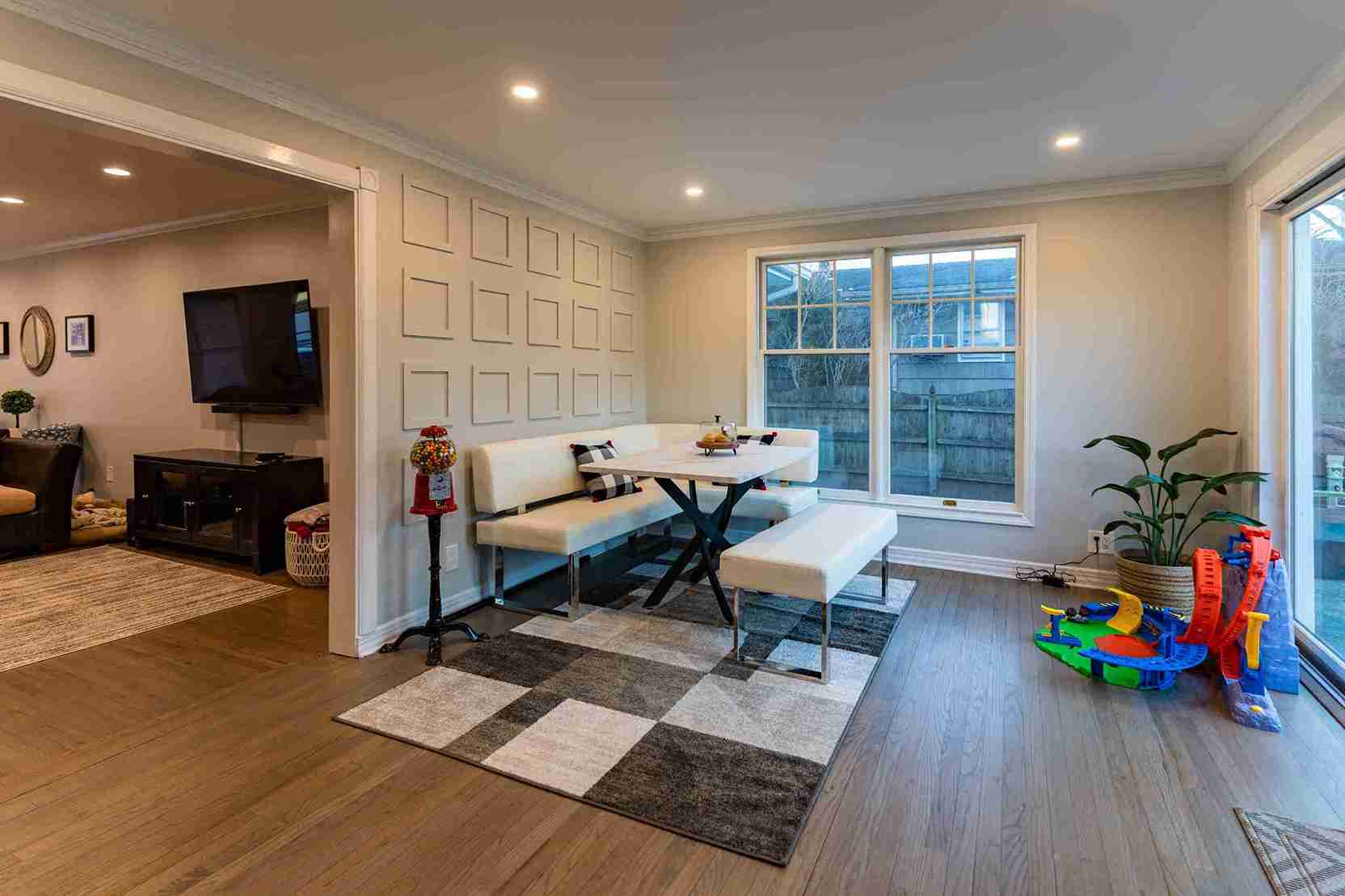 Cozy dining nook with white upholstered bench seating, geometric wall paneling, large windows, and children's toys neatly placed beside a potted plant on wood flooring.