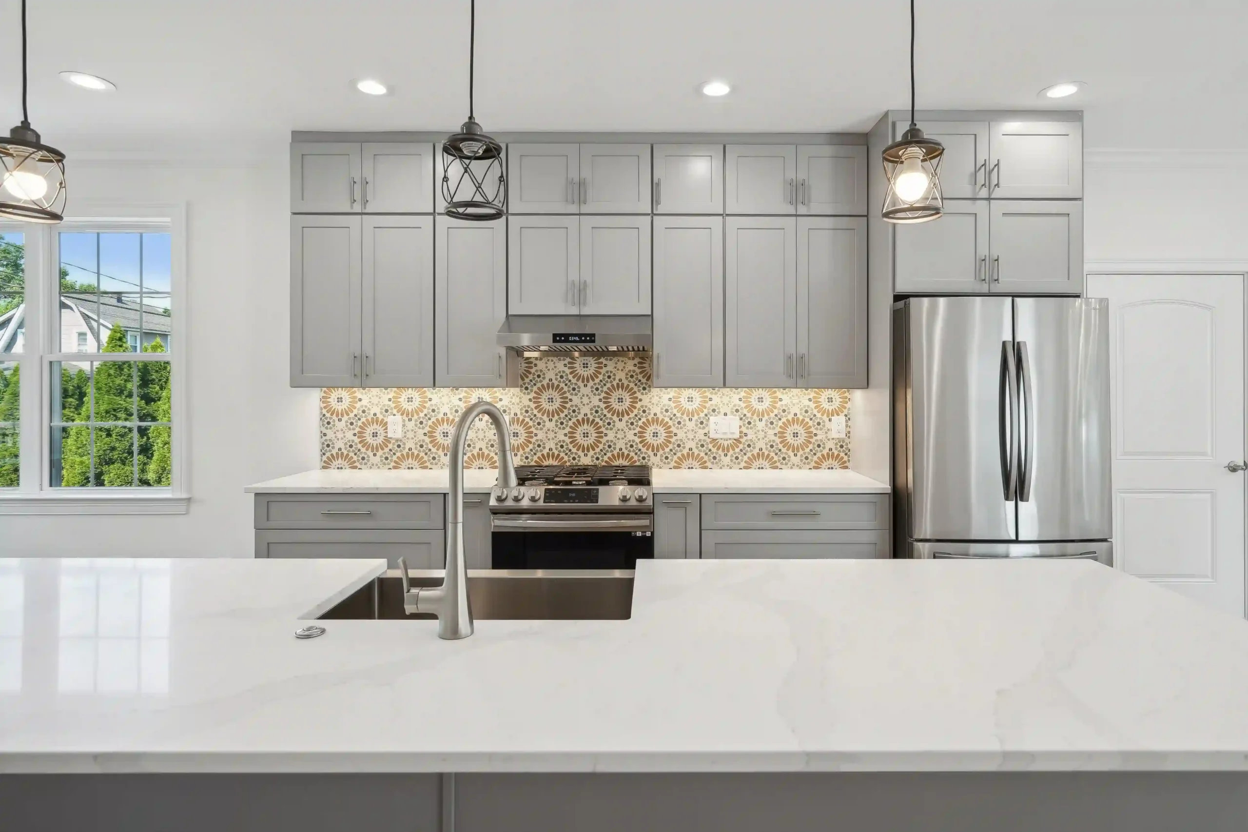 A modern kitchen featuring gray cabinets, a stainless steel refrigerator, and decorative tile backsplash with pendant lighting above an island with a sink.