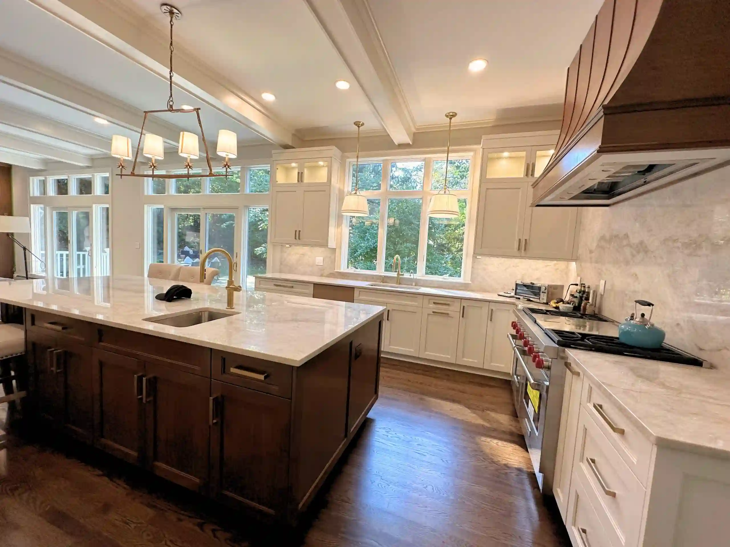 Luxury kitchen with a large marble island, white cabinetry, dark wood accents, coffered ceiling, pendant lighting, and expansive windows bringing in natural light.