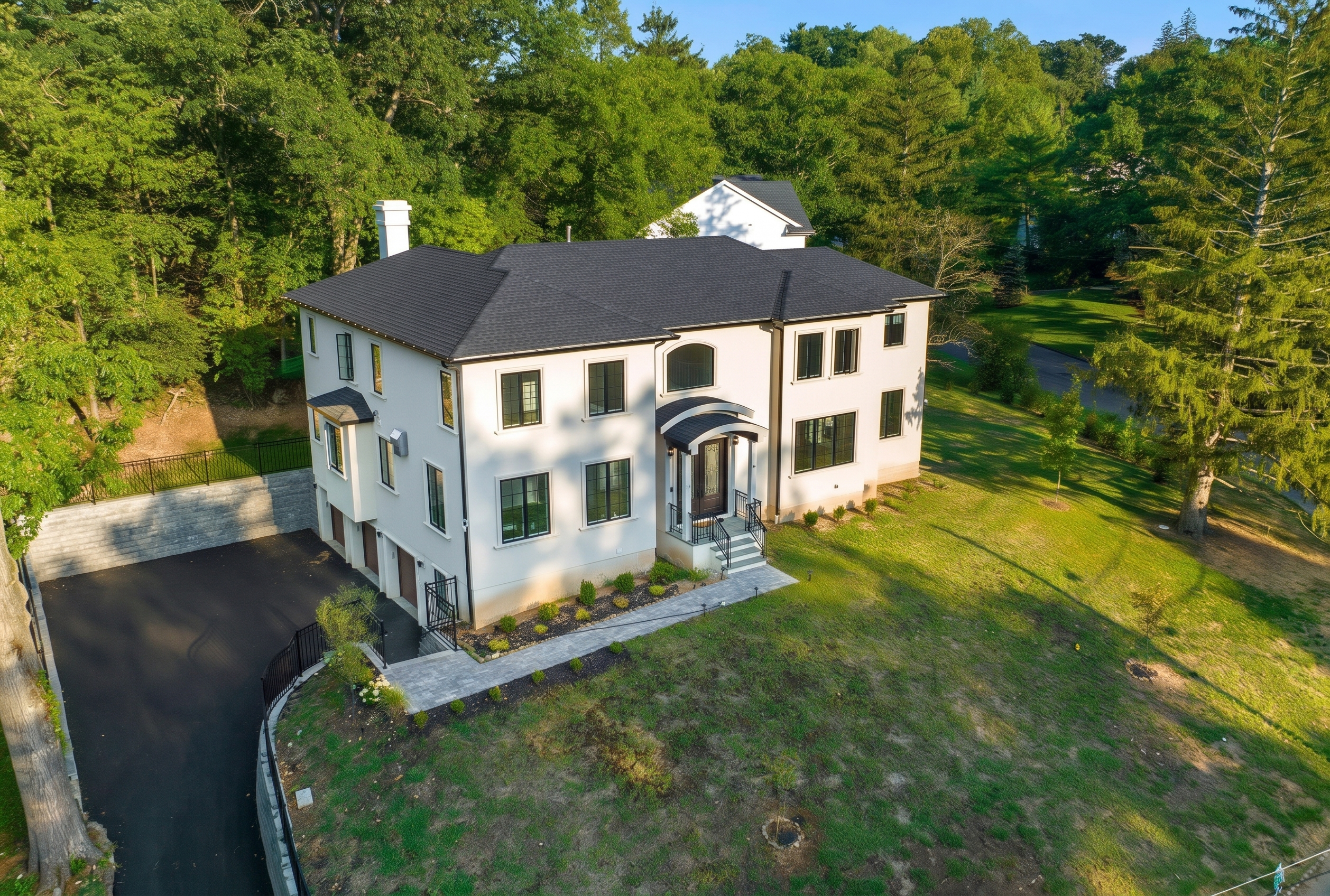Aerial view of a WA Construct custom home with white stucco exterior, black-framed windows, arched entry portico, tuck-under three-car garage, and paver walkway on a wooded lot.