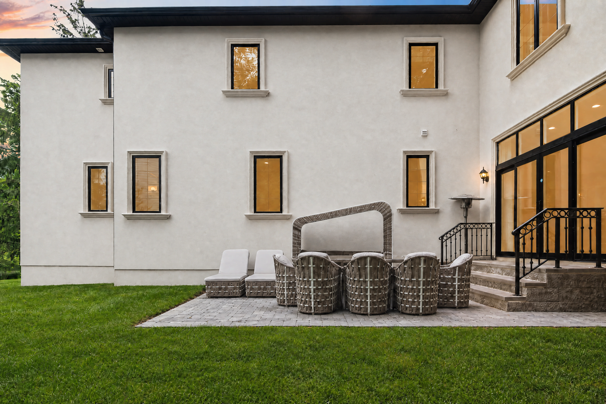 Backyard patio and rear elevation of a WA Construct custom home at dusk, with stucco exterior, cast stone window surrounds, a paver patio with woven outdoor seating, and wrought iron stair railings.