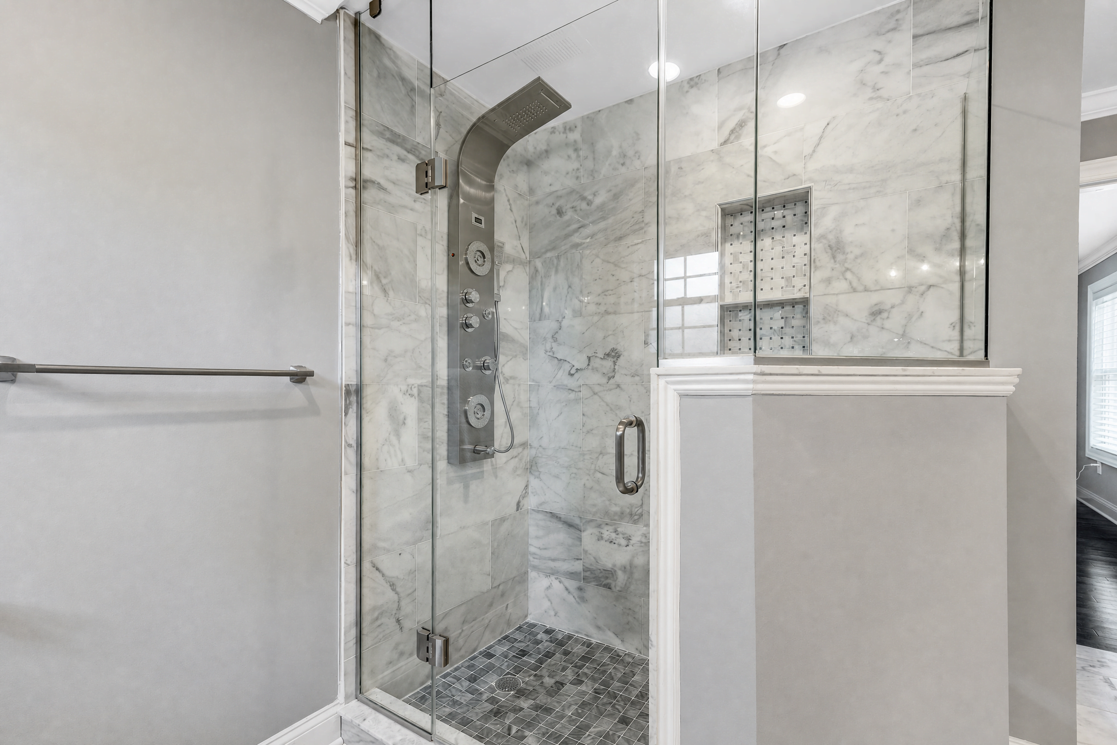 Custom walk-in shower by WA Construct with frameless glass enclosure, marble-look tile walls, a stainless steel multi-function shower panel, mosaic tile niche, and mosaic tile shower floor.