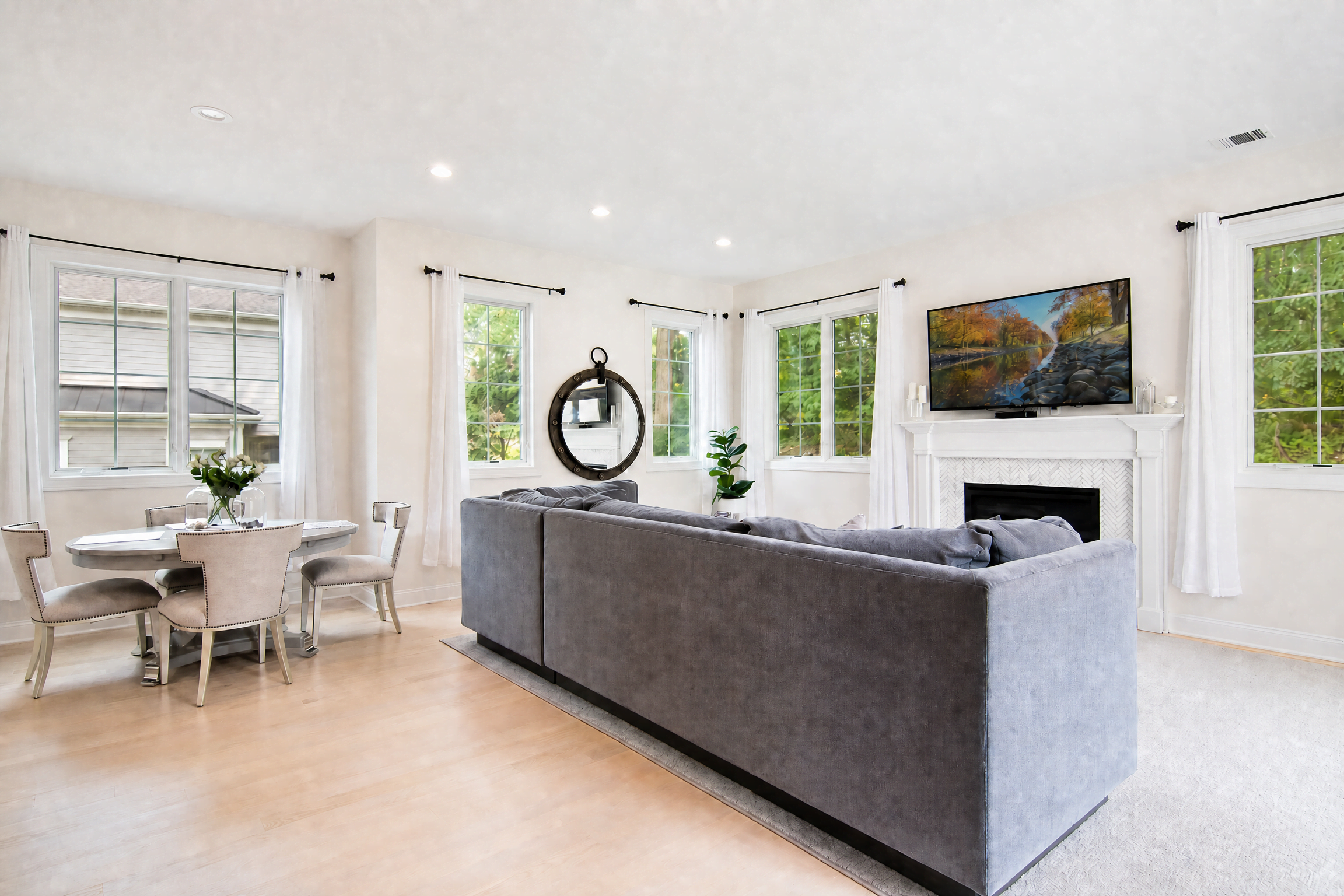 Family room in a WA Construct custom home with a gray sectional sofa, gas fireplace with white mantel and herringbone tile surround, adjoining breakfast dining area, and light hardwood flooring.