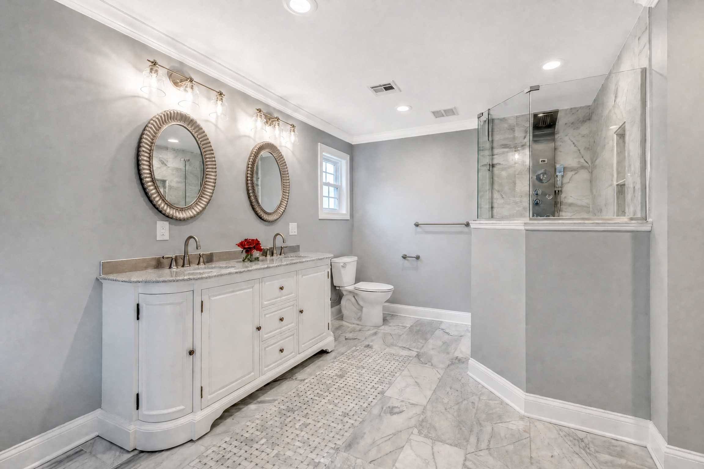 Primary bathroom by WA Construct with a white double vanity, granite countertop, oval framed mirrors, glass-enclosed walk-in shower, and marble tile flooring with a basketweave mosaic inlay.
